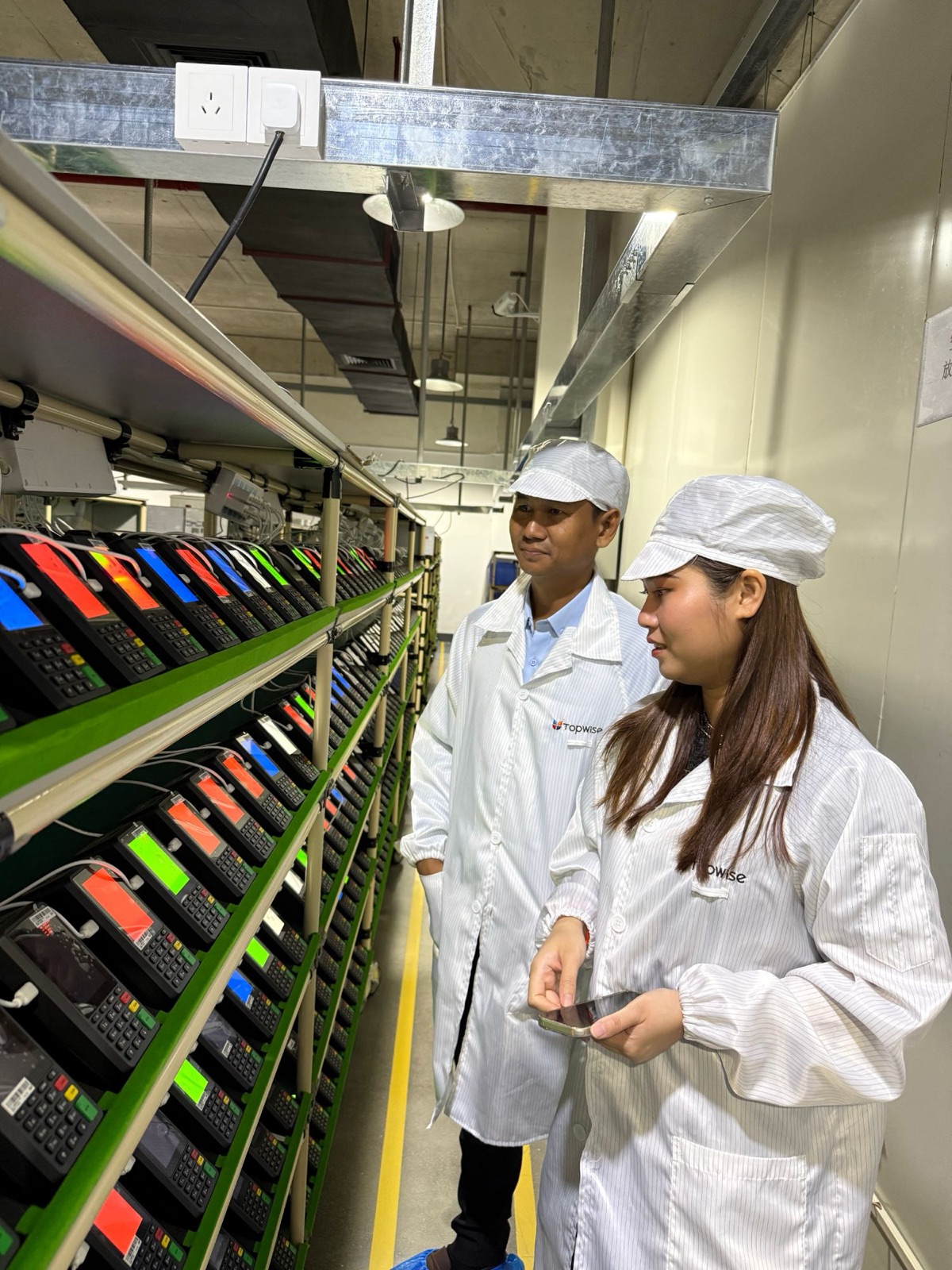 Two people in Topwise lab coats checking rows of payment terminals on a factory test line