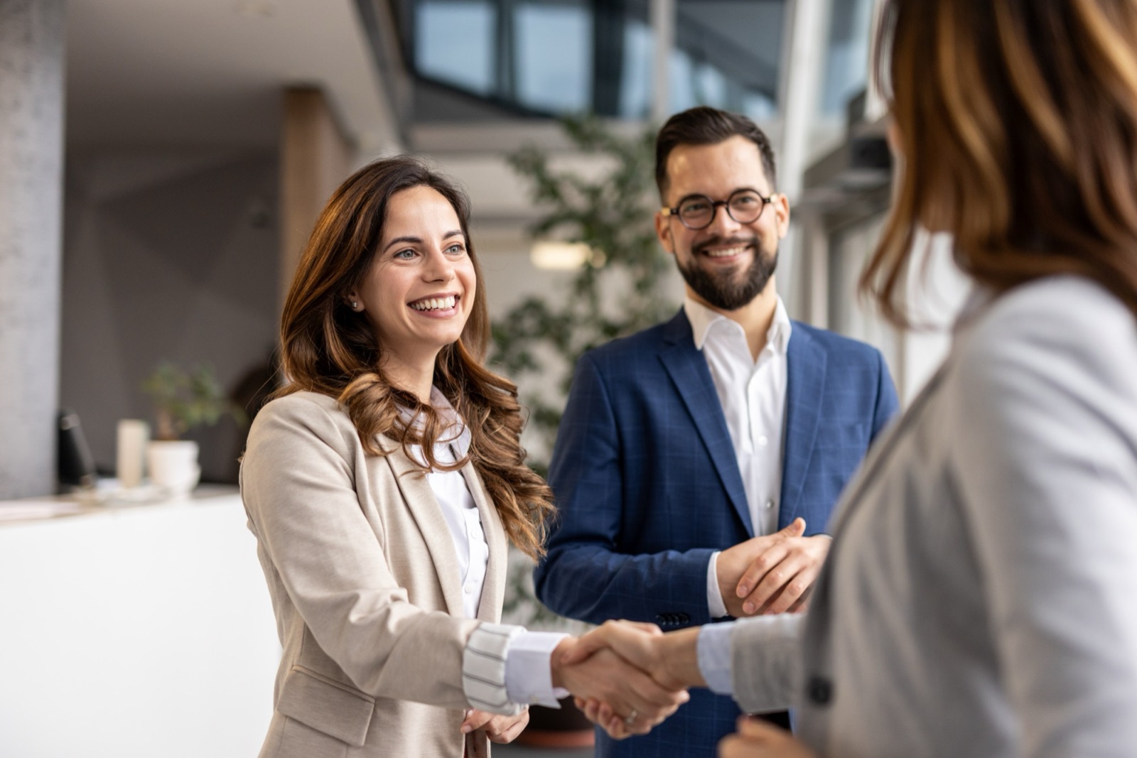 Two business partners shaking hands in a bright office lobby while a colleague looks on.