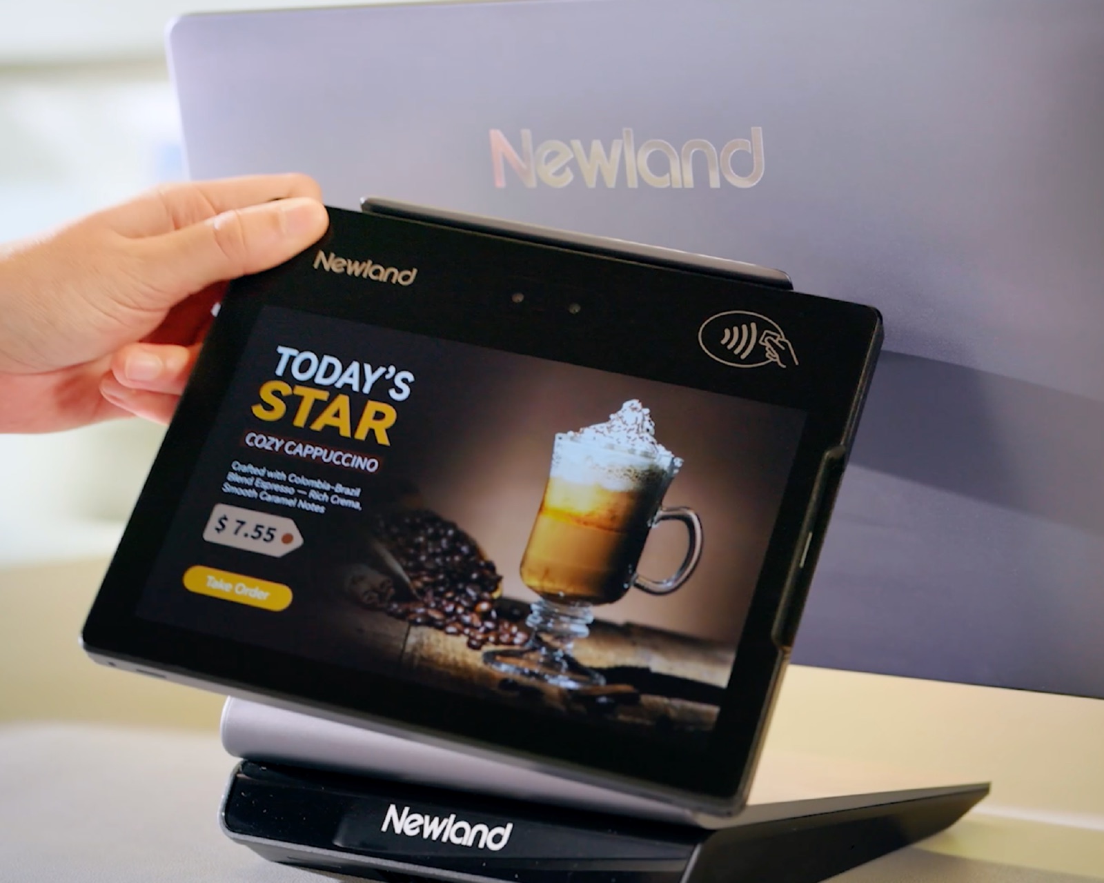 Hand holding a Newland POS customer display showing a café menu offer, with the main display behind